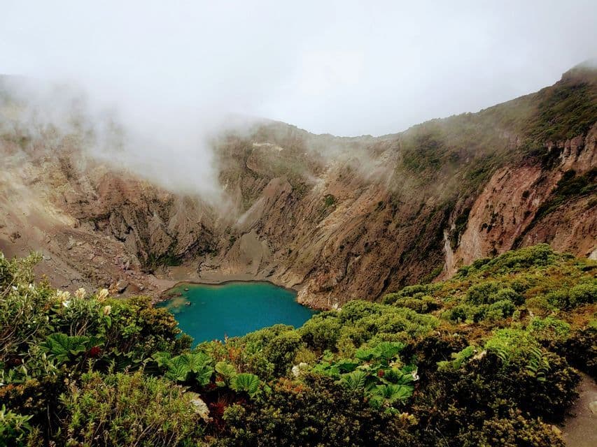 Vista aérea de un lago turquesa enclavado en un gran cráter volcánico, con follaje verde en primer plano y neblina en lo alto.