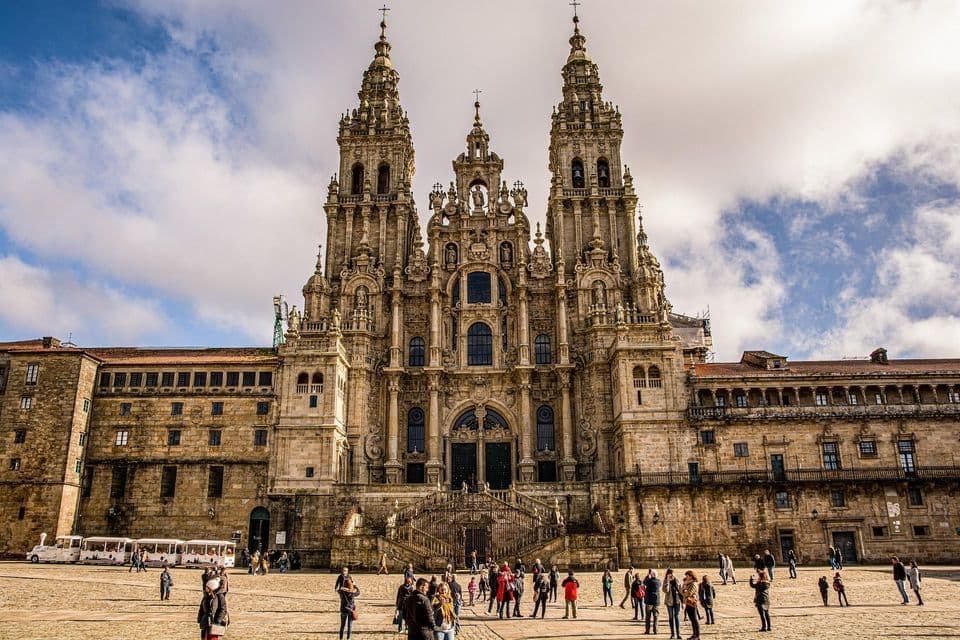 Una ornamentada catedral de piedra con campanarios gemelos se alza sobre una bulliciosa plaza llena de gente en un día parcialmente nublado.