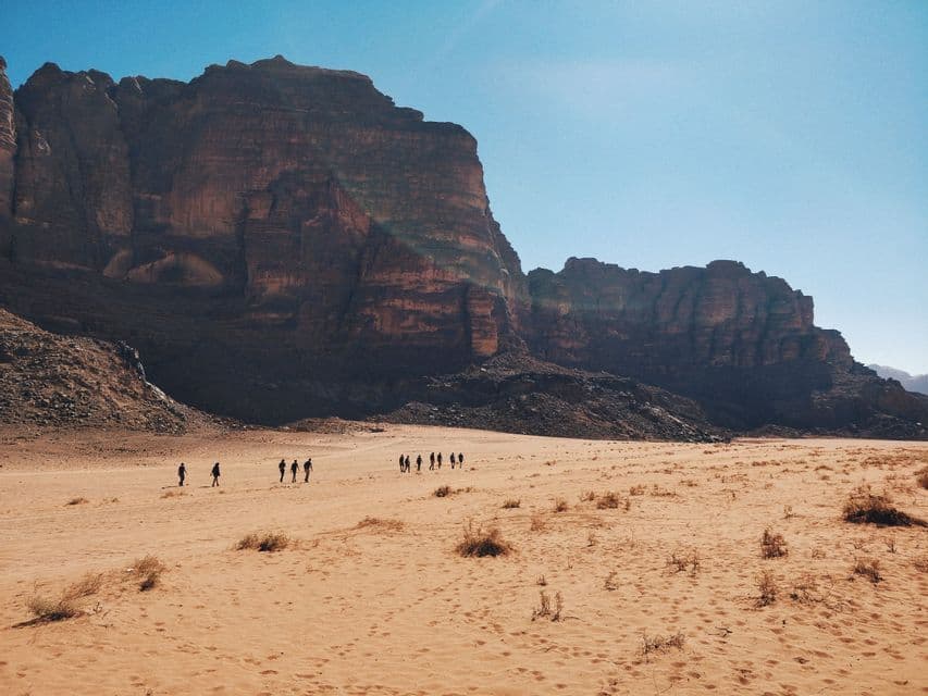 A WeRoad group trip walks across a vast desert landscape with large rock formations in the background under a blue sky.