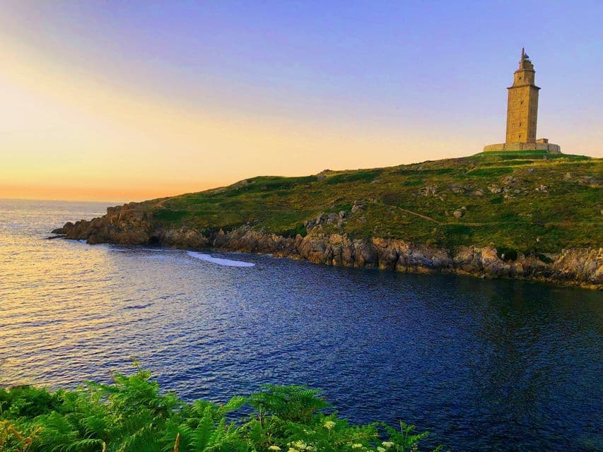 Una torre de faro de piedra se alza sobre una colina cubierta de hierba en una costa rocosa, con vistas al océano en calma durante el atardecer.