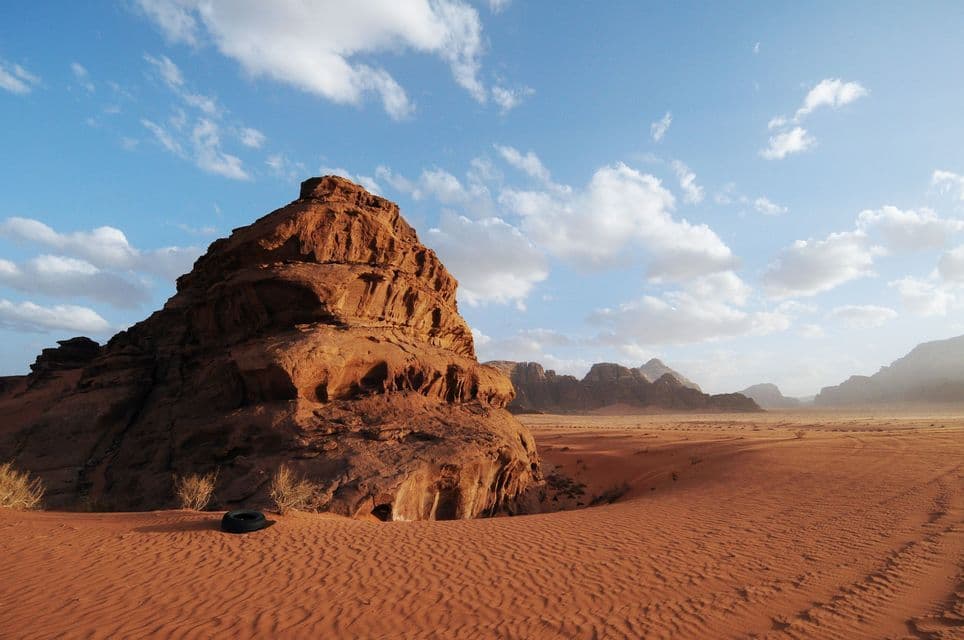 A large, red rock formation rises from a desert of rippled sand under a blue sky with white clouds, and a black tire rests in the foreground.