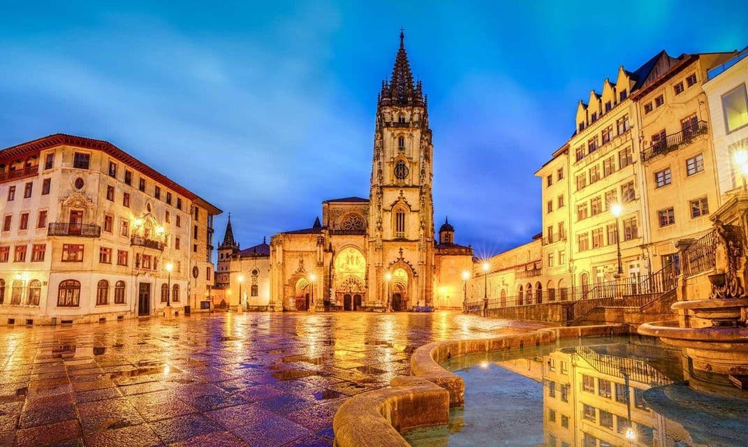 Una gran catedral iluminada se alza en una plaza pública mojada al anochecer, sus luces reflejándose en los adoquines.