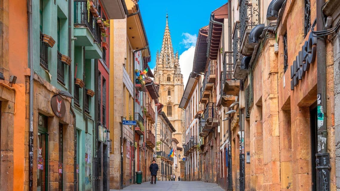 Una calle estrecha con edificios coloridos conduce hacia la alta y ornamentada aguja de una catedral bajo un cielo azul brillante.