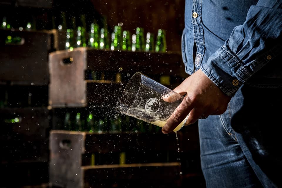 Una persona con camisa de mezclilla sostiene un vaso mientras una bebida amarilla burbujeante salpica, con botellas verdes borrosas al fondo.
