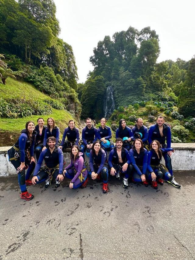 Un viaje en grupo de WeRoad con trajes de neopreno y arneses posando para una foto en una carretera con una cascada y un bosque exuberante al fondo.