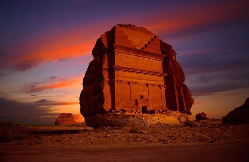 Una grande tomba scolpita in una formazione rocciosa in un paesaggio desertico, illuminata dai colori di un tramonto drammatico.