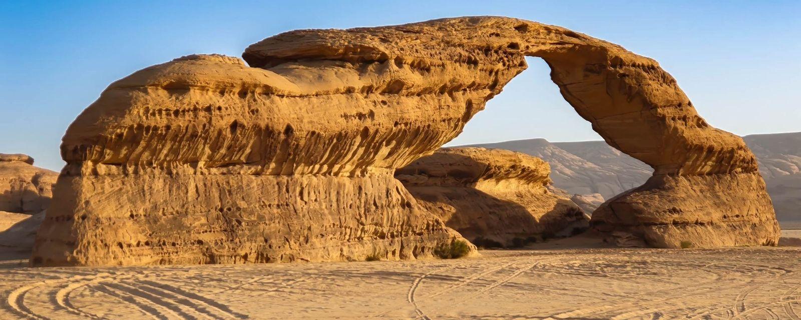 Una grande formazione rocciosa di arenaria che forma un arco naturale in un paesaggio desertico sotto un cielo azzurro.