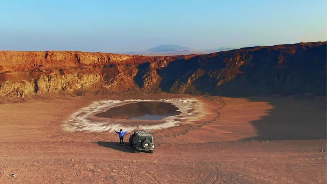 Una persona è in piedi con le braccia distese accanto a un veicolo fuoristrada, affacciandosi su un cratere desertico con un piccolo lago al centro.