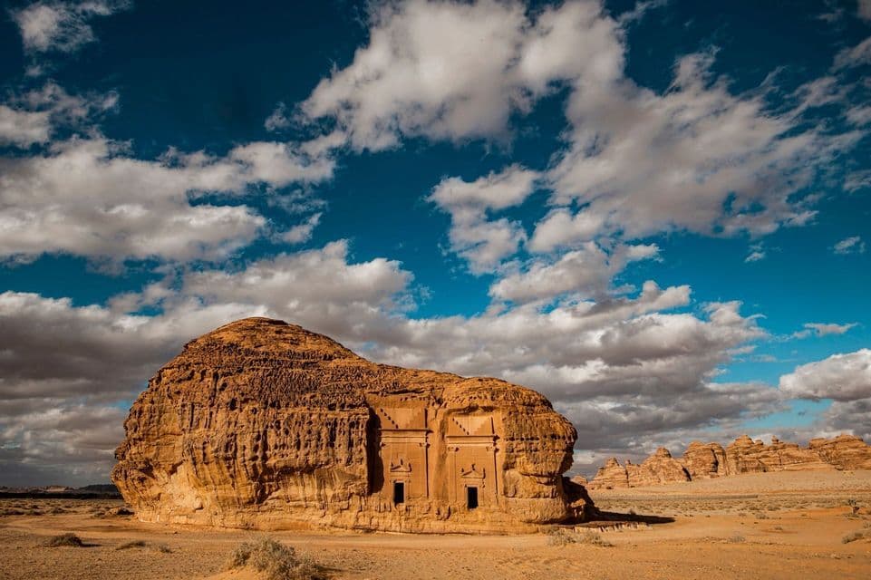 Un'ampia formazione rocciosa in arenaria con una facciata scolpita con due ingressi si trova in un deserto sotto un cielo azzurro nuvoloso.