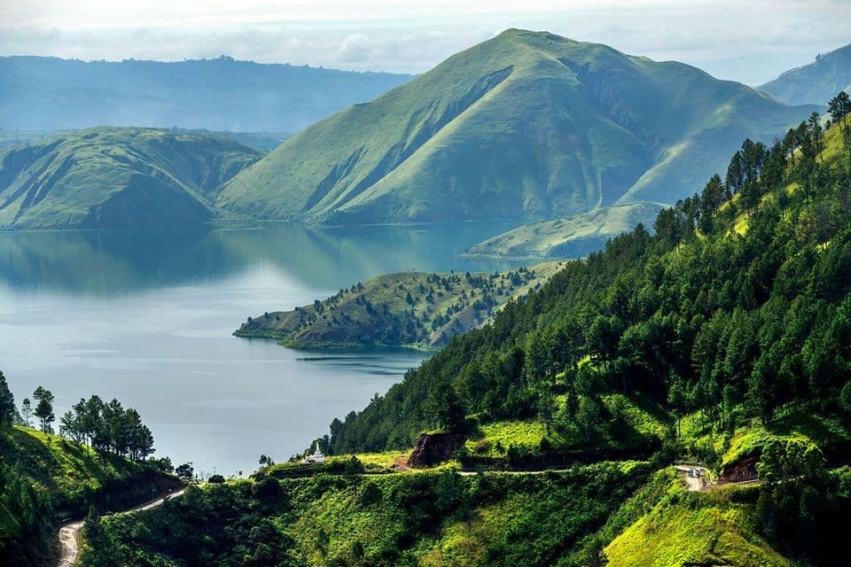 Una vista dall'alto di un vasto lago circondato da dolci montagne verdi, con una strada tortuosa che attraversa i fianchi delle colline boscose.