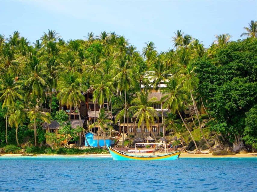 Una barca colorata galleggia su acqua blu di fronte a una spiaggia sabbiosa costeggiata da fitte palme e edifici.