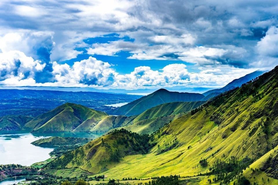Una vista panoramica di lussureggianti colline verdi e montagne che circondano un grande lago sotto un cielo nuvoloso e drammatico.