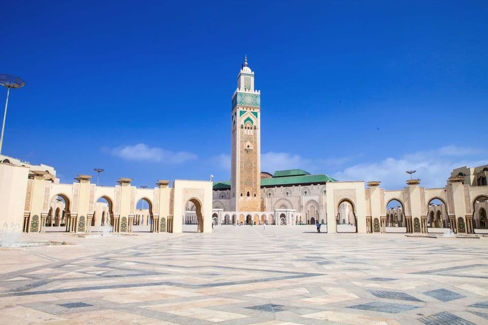 Una gran mezquita con un minarete alto y ornamentado se alza tras una amplia plaza con motivos y arcos, bajo un cielo azul claro.