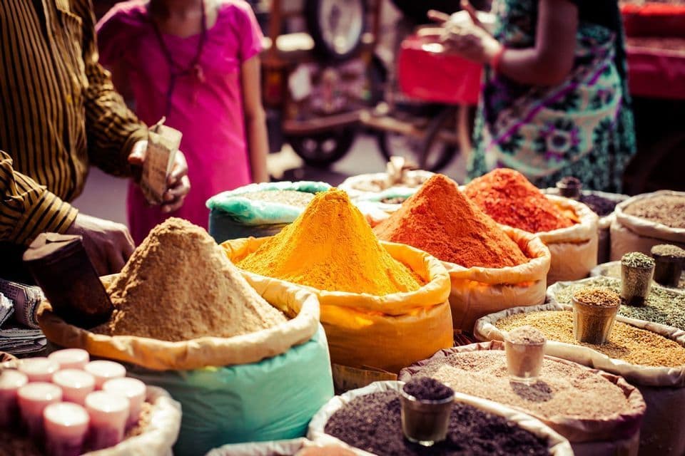 Colorful piles of ground spices like turmeric and chili powder displayed in large sacks at a busy outdoor market stall.