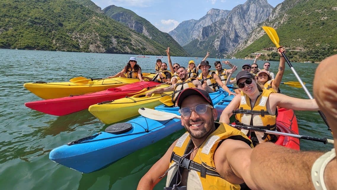Un groupe WeRoad prend un selfie en faisant du kayak ensemble sur un grand lac entouré de montagnes.