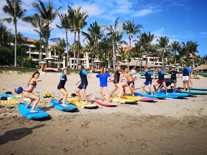 A WeRoad group trip taking a surfing lesson on a sandy beach, practicing on colorful surfboards under palm trees.