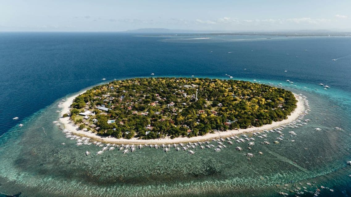Una vista aérea de una pequeña y exuberante isla tropical con una playa de arena blanca, rodeada de aguas cristalinas de color turquesa y muchos barcos anclados.