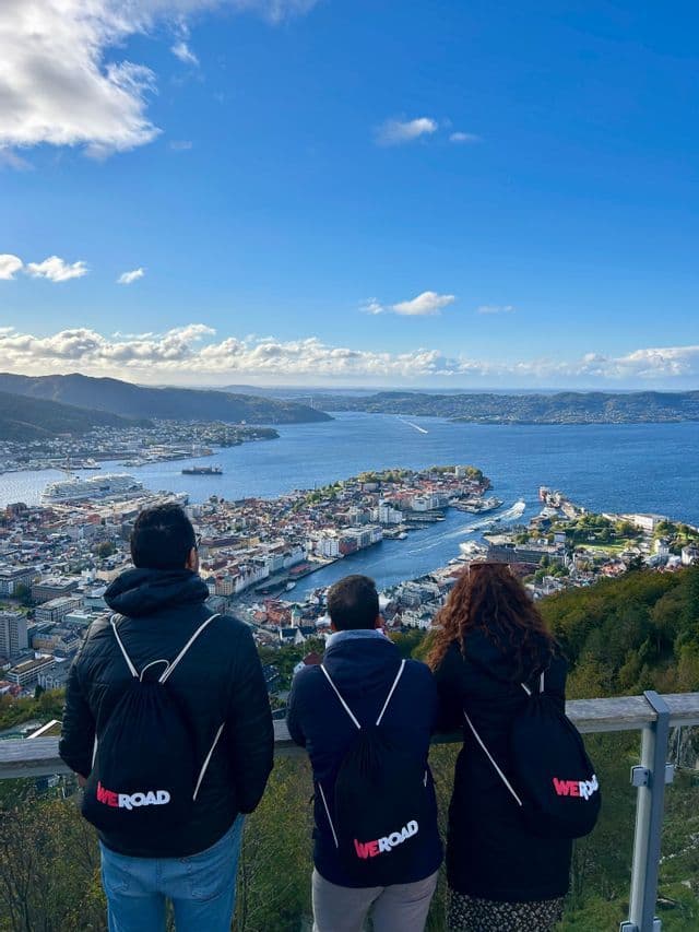 Un gruppo WeRoad di tre persone, viste di spalle, che ammirano un panorama su una città costiera e una baia da un punto elevato.