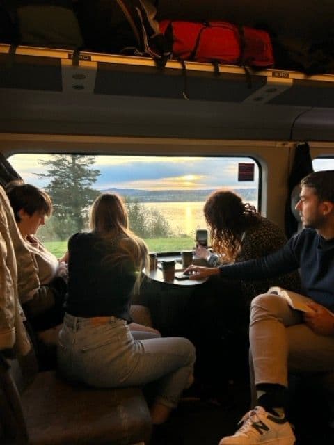 Un viaggio di gruppo WeRoad, seduti attorno a un tavolo su un treno, mentre si guarda fuori dal finestrino un tramonto su uno specchio d'acqua.