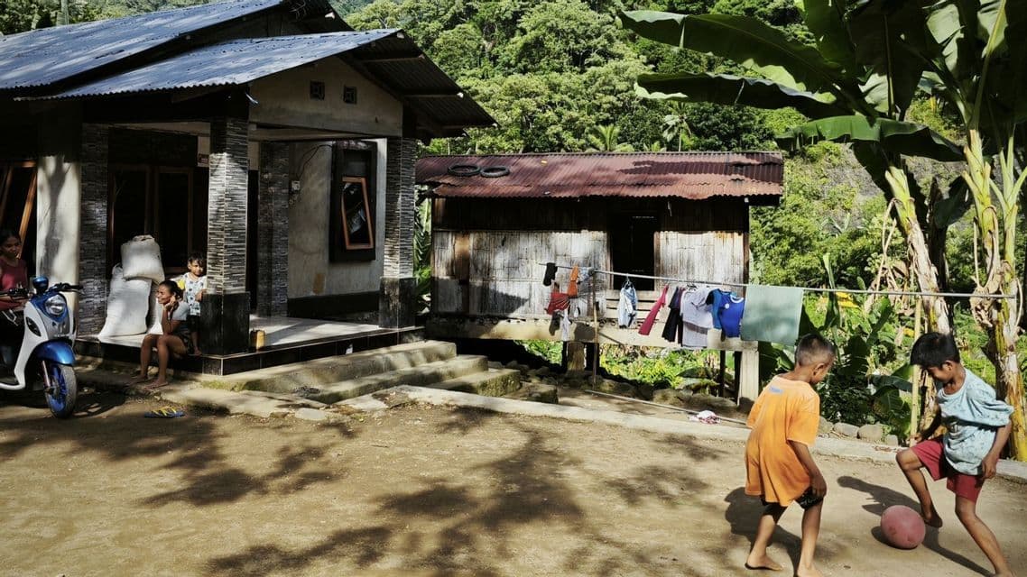 In einem ländlichen Dorf spielen zwei barfüßige Jungen auf einem staubigen Hof Fußball, während andere auf einer Veranda sitzen.