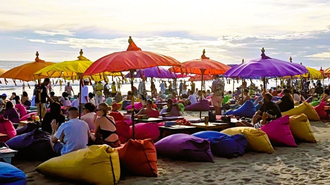 Eine große Menschenmenge entspannt sich auf farbigen Sitzsäcken unter bunten Sonnenschirmen an einem Sandstrand.