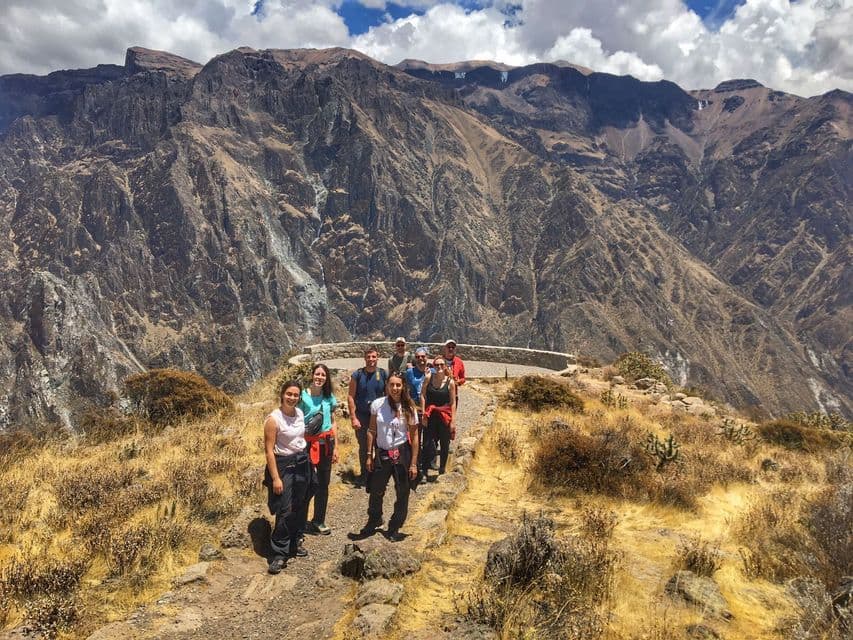 Un gruppo WeRoad in viaggio sorride alla fotocamera su un sentiero di montagna polveroso, con un grande canyon roccioso che si estende dietro di loro.