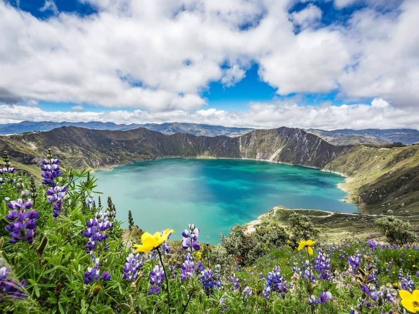 A turquoise crater lake surrounded by mountains, with purple and yellow wildflowers in the foreground under a cloudy blue sky.