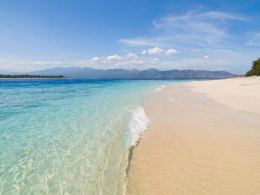 Clear turquoise water gently laps onto a white sand beach with mountains in the background under a blue sky.