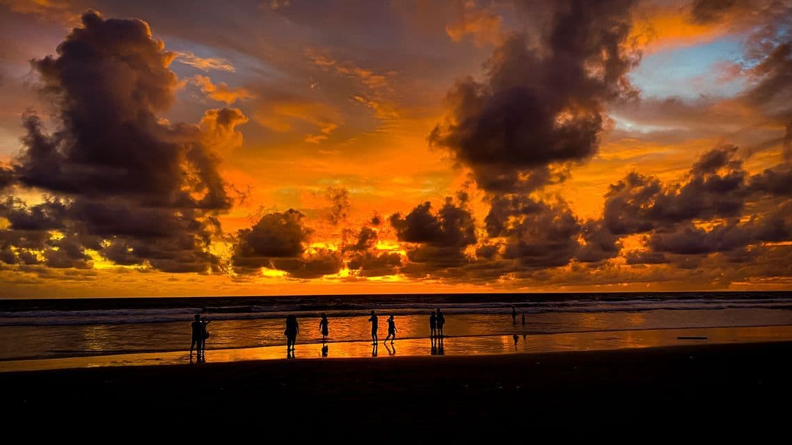 Un groupe WeRoad en voyage, silhouetté sur une plage humide, observant un coucher de soleil orange spectaculaire avec des nuages sombres au-dessus de l'océan.