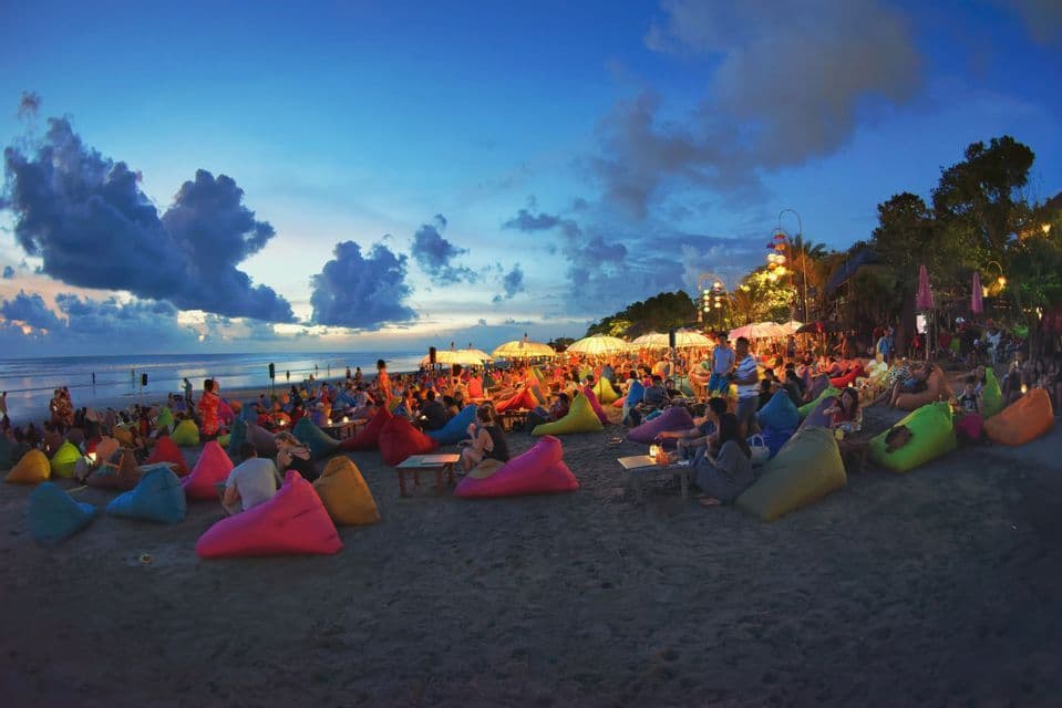 A WeRoad group trip relaxes on colorful beanbags on a sandy beach at twilight, with illuminated umbrellas overhead.