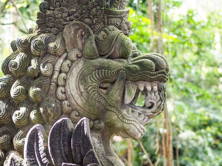 A close-up profile of an intricate, moss-covered stone sculpture of a mythical creature with fangs, set in a green jungle.