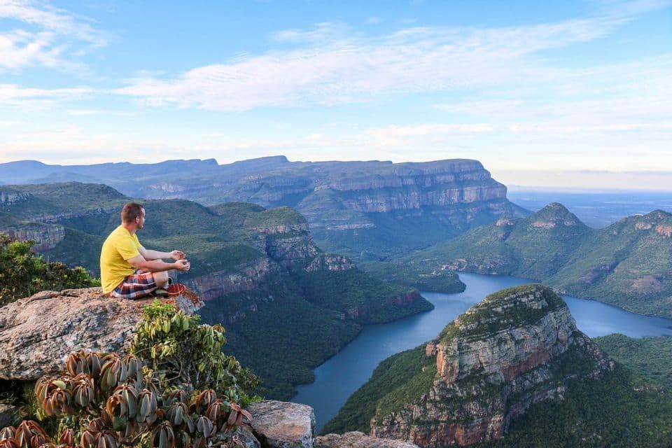 A man in a yellow t-shirt sits cross-legged on a rocky cliff, looking out over a vast green canyon and a winding river below.