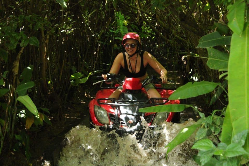 Eine Frau mit rotem Helm fährt ein rotes Quad und spritzt durch Wasser in einem dichten, sonnendurchfluteten Dschungel.