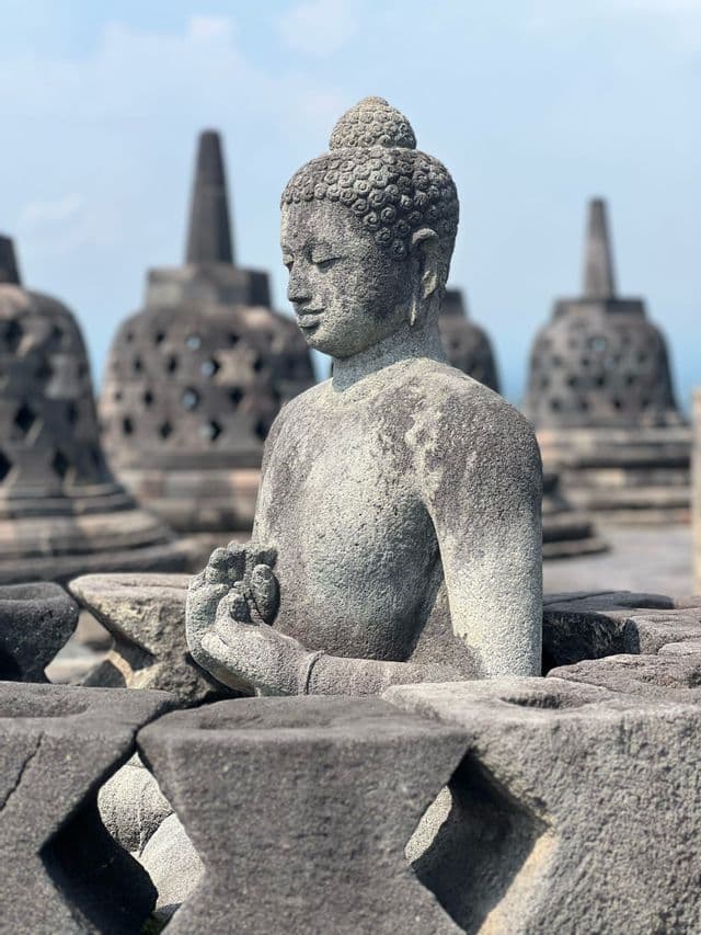 Eine verwitterte Stein-Buddha-Statue sitzt in meditativer Pose, mit glockenförmigen Stupas im Hintergrund vor klarem Himmel.