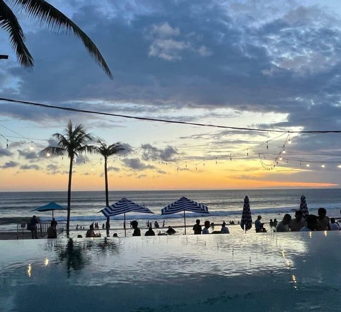 Silhouetten einer WeRoad-Gruppenreise in einem Infinity-Pool bei einem bewölkten Sonnenuntergang über dem Meer, mit Palmen und Lichterketten.