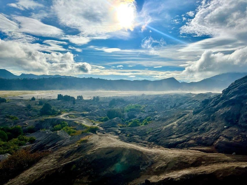 Eine weite Vulkanlandschaft mit dunklem, felsigem Gelände und spärlicher Vegetation unter einem hellen, sonnigen Himmel mit Wolken.