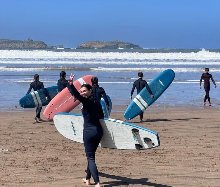 Eine WeRoad Gruppenreise in Neoprenanzügen mit Surfbrettern an einem Sandstrand, während eine Frau vorne lächelnd ein Friedenszeichen macht.