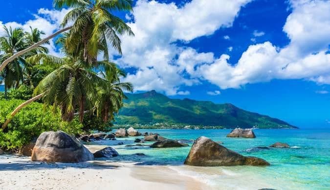Palme su una spiaggia di sabbia bianca con grandi rocce, acqua turchese e una montagna verde sullo sfondo sotto un cielo parzialmente nuvoloso.