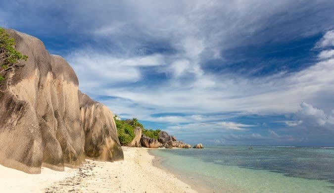 Grandi massi di granito giacciono su una spiaggia di sabbia bianca, accanto ad acque cristalline turchesi, sotto un cielo azzurro con nuvole bianche.