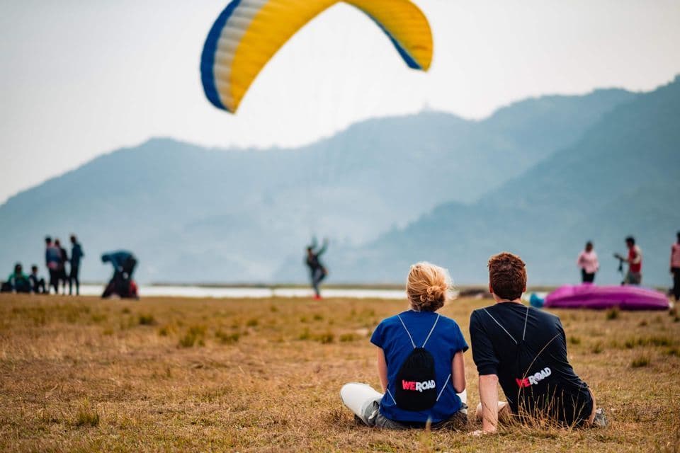 Dos personas de un viaje grupal de WeRoad se sientan en un campo de hierba, observando un parapente amarillo y azul volar sobre montañas lejanas.