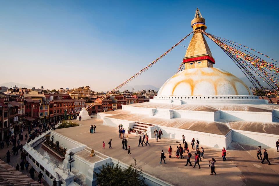 A high-angle view of people walking on the terraces of a large white Buddhist stupa with a golden spire and colorful prayer flags.