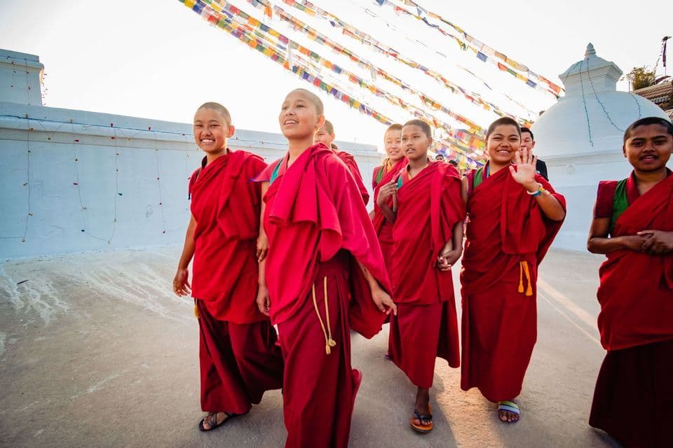 Un groupe de jeunes nonnes bouddhistes en robes rouges souriantes et marchant devant un stupa blanc sous des drapeaux de prière colorés.