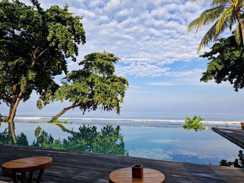 Ein Infinity-Pool auf einem Holzdeck mit Blick auf den Ozean, der Bäume und einen teilweise bewölkten blauen Himmel spiegelt.