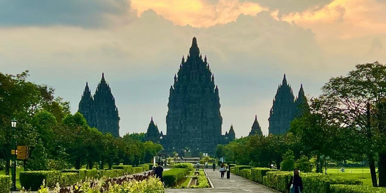 Ein weiter Blick entlang eines gepflasterten Parkwegs zu einem kunstvollen Steintempelkomplex mit mehreren Türmen unter bewölktem Himmel.