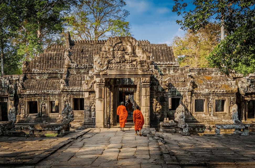 Zwei Mönche in orangefarbenen Roben gehen einen Steinpfad entlang zum Eingang eines alten, reich verzierten Tempels, der von Bäumen umgeben ist.