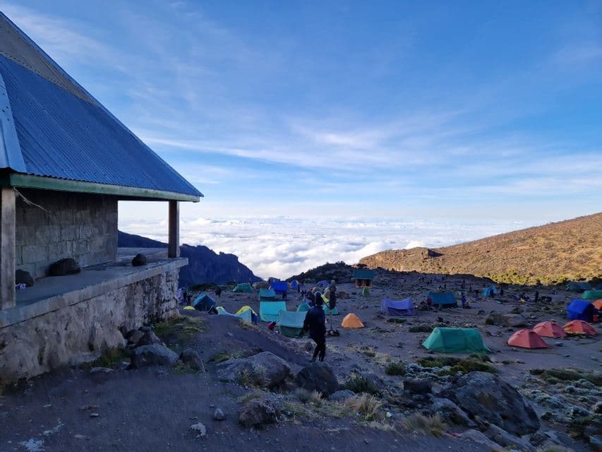 A mountain base camp with a stone building and colorful tents spread across a rocky landscape above the clouds.