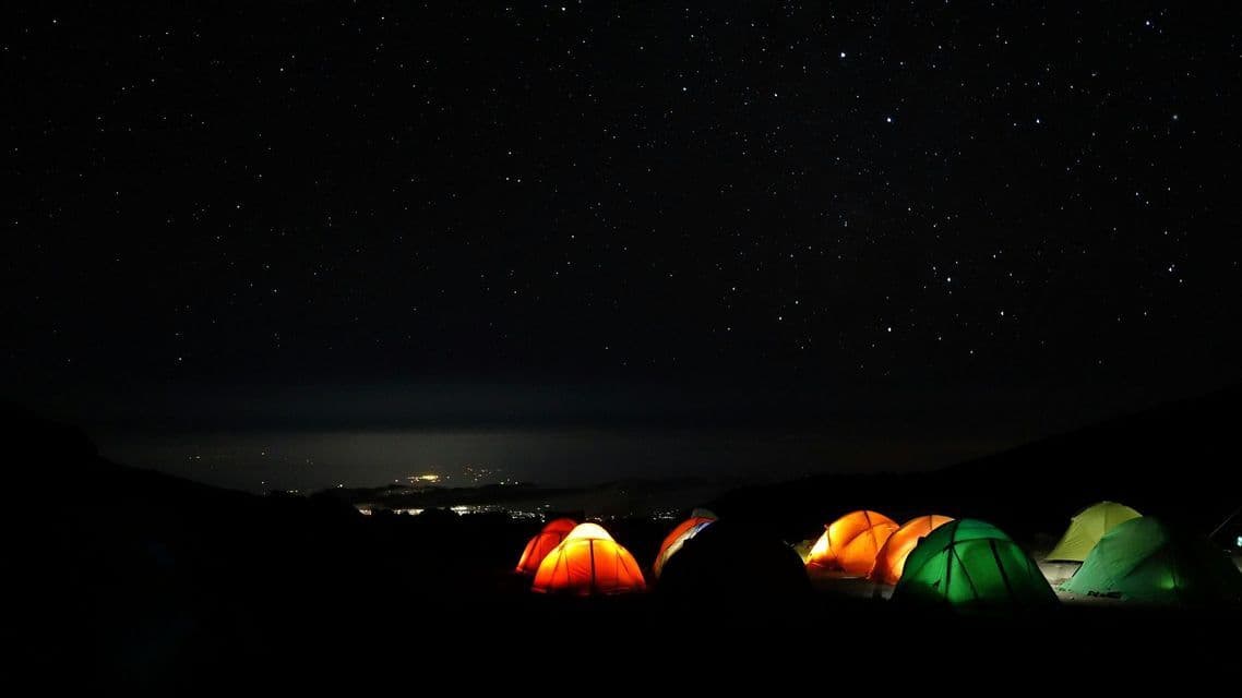 Several illuminated tents glow at a campsite under a starry night sky, with distant city lights visible below.