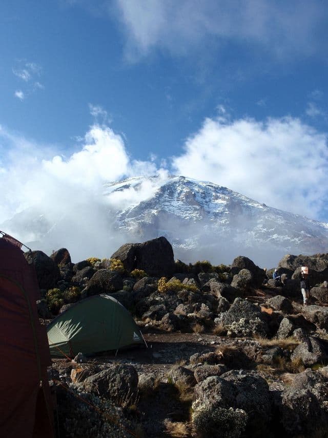 Tents pitched on a rocky campsite with a large, snow-capped mountain looming in the background under a blue sky with clouds.