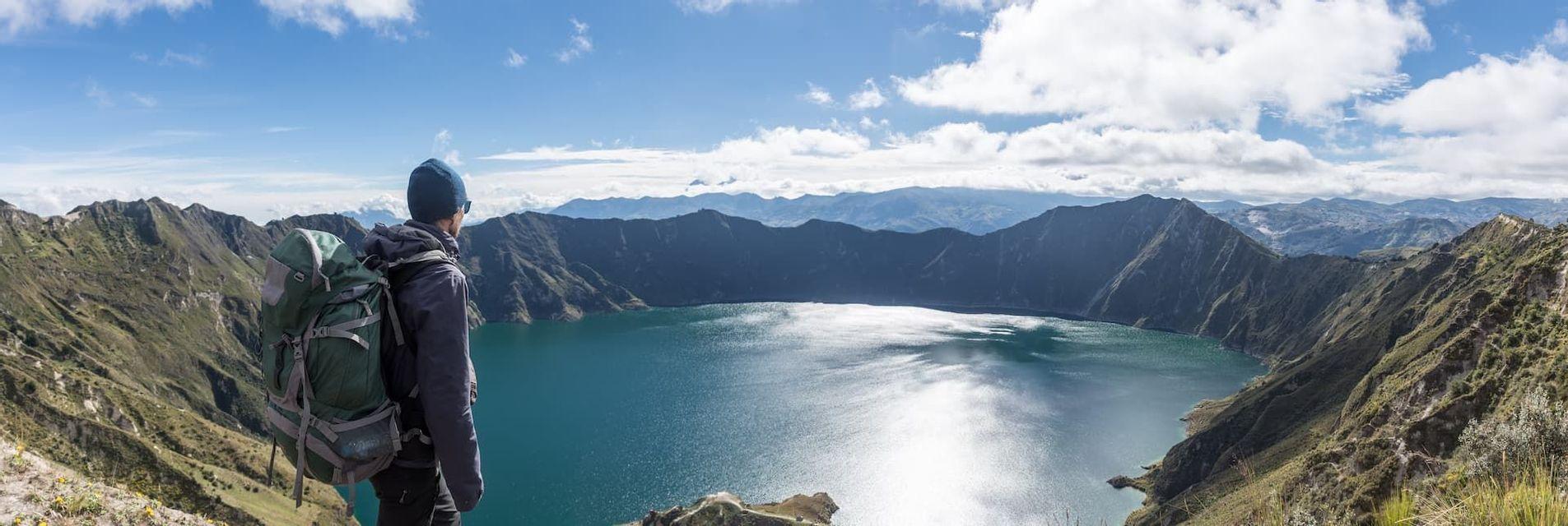 Un randonneur avec un sac à dos se tient sur une crête de montagne, surplombant un vaste lac de cratère sous un ciel bleu partiellement nuageux.