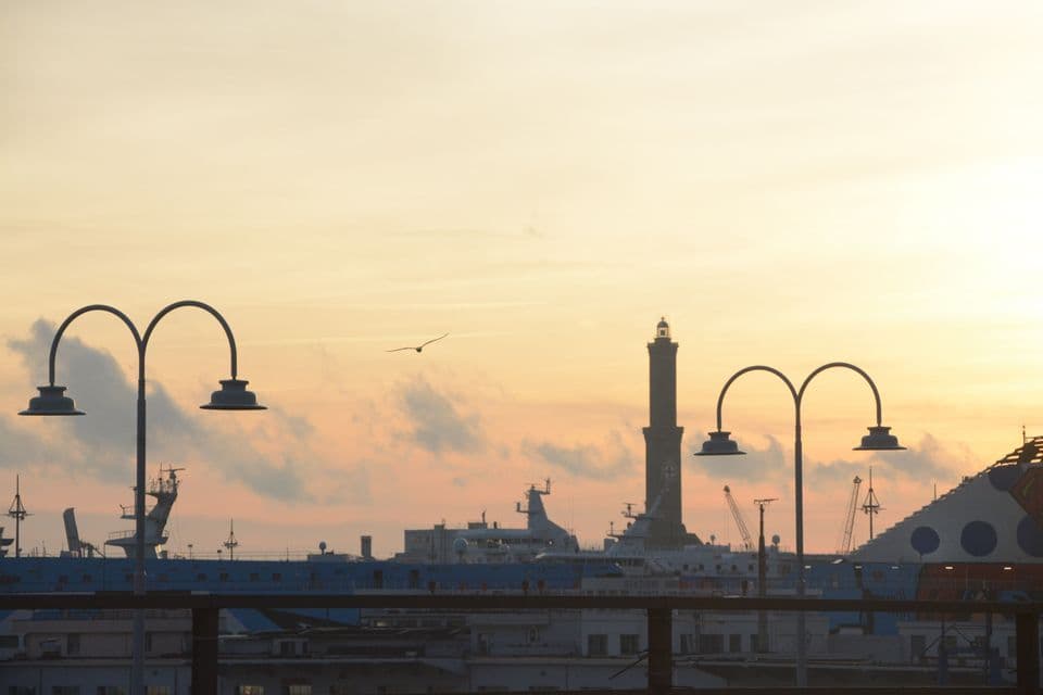 Un faro e lampioni si stagliano su un porto con navi e gru sotto un cielo caldo al crepuscolo.